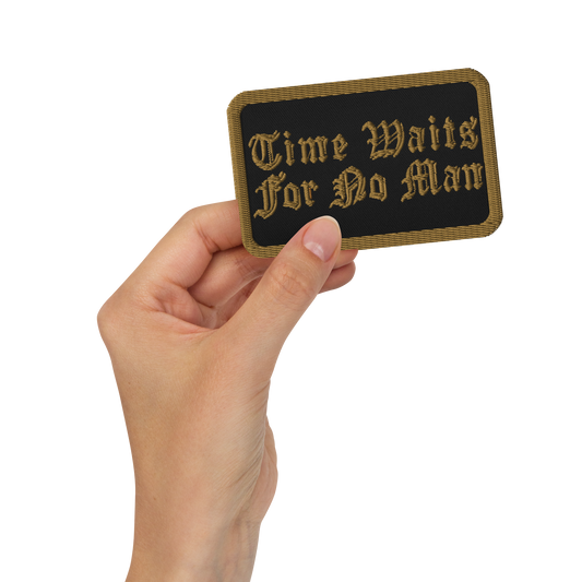 A hand holds the Mainstay Entertainment Time Waits For No Man "Logo" Embroidered Patch—rectangular, black, with gold trim and ornate text reading “Time Waits For No Man.”.