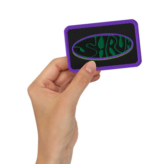 A hand holds the SHRÜM "Logo" Embroidered Patch by Mainstay Entertainment—a black patch with a purple border, green and purple oval design, and stylized SHRÜM text. Ideal as an iron-on patch for jackets, bags, or hats.