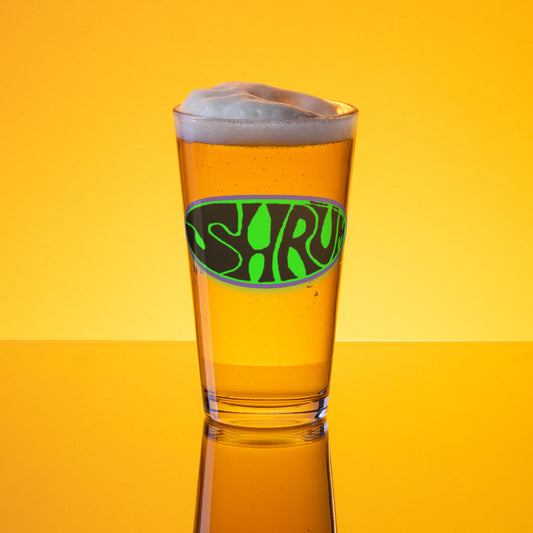 A SHRÜM "Logo" Pint Glass by Mainstay Entertainment, filled with golden beer and a foamy head, displays the green and black logo against a vibrant orange-yellow background with reflections on the surface.
