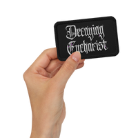 A hand holds the Mainstay Entertainment Decaying Eucharist "Logo" embroidered patch, featuring silver gothic lettering on a black rectangle, against a plain white background.