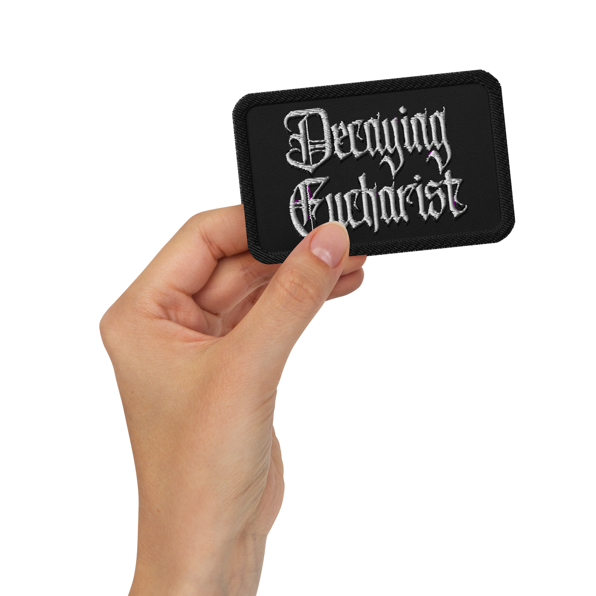 A hand holds the Mainstay Entertainment Decaying Eucharist "Logo" embroidered patch, featuring silver gothic lettering on a black rectangle, against a plain white background.