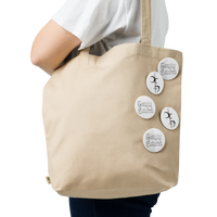 A person carries a beige tote bag decorated with Mainstay Entertainment’s Decaying Eucharist "Logo" Set of 5 buttons, each with black handwritten-style designs, attached to the side, while wearing a white shirt.