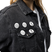 A person wears a black denim jacket and white shirt, displaying Mainstay Entertainment’s Decaying Eucharist "Logo" Set of (5) scratch and UV-resistant pin buttons with black designs and text on the jacket’s upper left side and pocket flap.