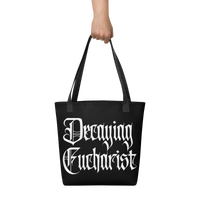 A hand holds the Decaying Eucharist "Logo" Tote Bag by Mainstay Entertainment, featuring large white gothic lettering.