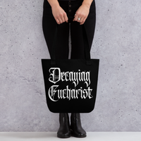 A person dressed in black holds the Mainstay Entertainment Decaying Eucharist "Logo" Tote Bag with white gothic lettering, standing against a light gray textured wall.