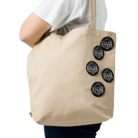 A person in a white shirt carries a beige tote bag adorned with Mainstay Entertainment's False Gods "Snake Logo" Set of 5 glossy buttons. The background is plain white.