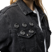 A person wears a black denim jacket and white shirt, accessorized with four Mainstay Entertainment False Gods "Snake Logo" buttons. The glossy buttons feature white text. Only the lower face and torso are shown.