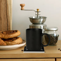 A black mug rests on a Mainstay Entertainment Flesh Parade "Classic Logo" ceramic coaster beside pastries, a manual coffee grinder, and a glass jar of coffee beans on a wooden surface.