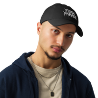 A young man wears the Flesh Parade "Embroidered Logo" Under Armour® Hat in black, paired with a beige t-shirt, blue zip-up hoodie, and layered silver necklaces as he looks at the camera against a plain backdrop.