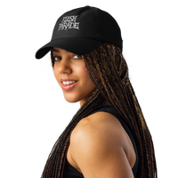 A woman with long braided hair wears the Flesh Parade "Embroidered Logo" Under Armour® Hat, featuring white text and a HeatGear sweatband, along with a black sleeveless top. She smiles while looking over her shoulder against a white background.