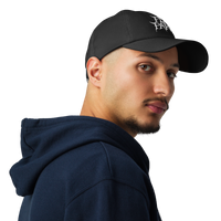 A young man with a short beard, wearing a Flesh Parade "Embroidered Logo" Under Armour® Hat and a dark blue hoodie, looks over his shoulder against a plain white background.