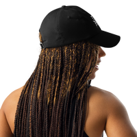 A woman with long braids wears the Flesh Parade "Embroidered Logo" Under Armour® Hat in black and a sleeveless top, seen from the back and slightly to the side against a plain white background.