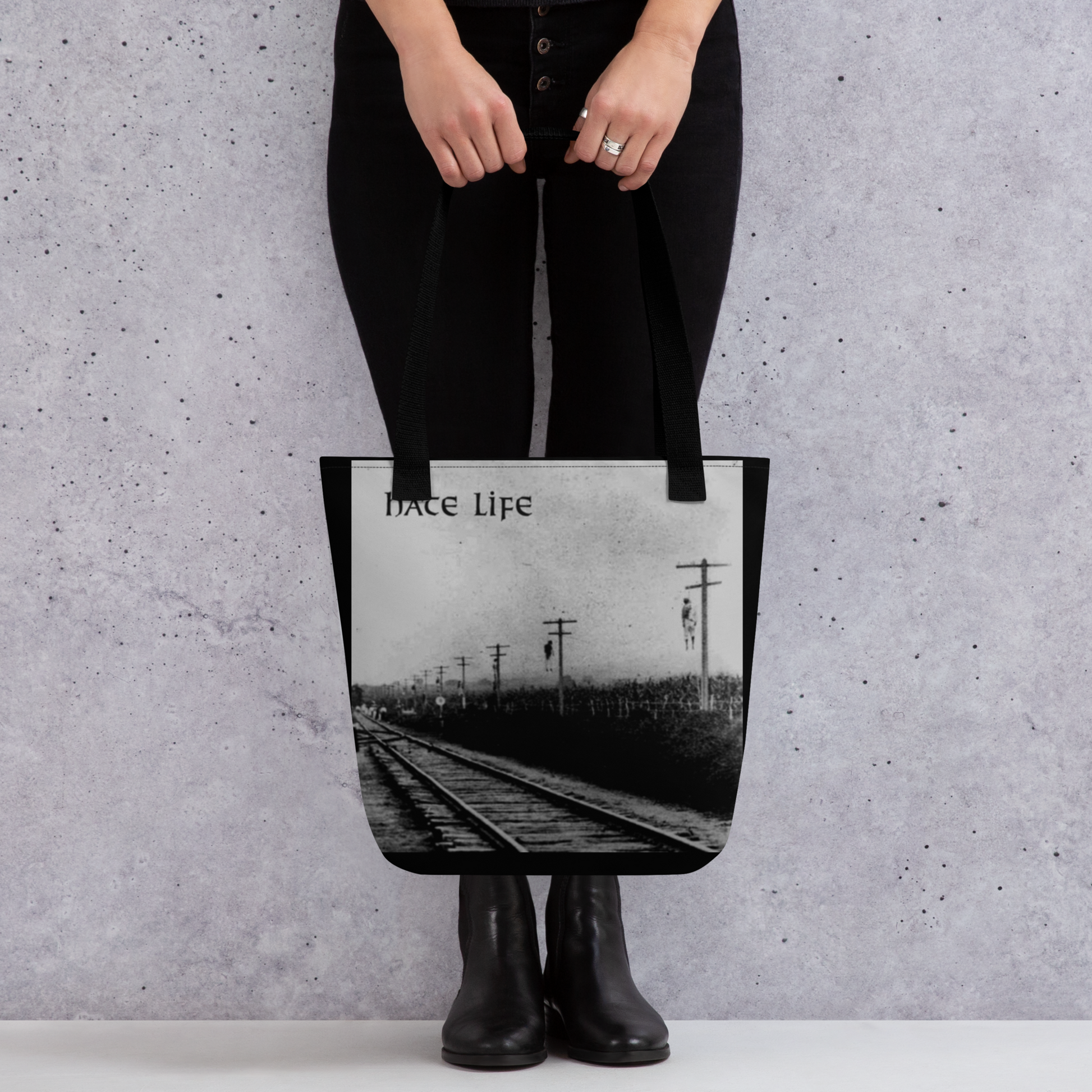 A person in black pants and boots holds the Mainstay Entertainment Flesh Parade "Hate Life" Tote Bag, featuring a black-and-white railroad track design with "hate Life" at the top, against a light textured wall.