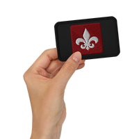 A hand holds the Mainstay Entertainment "Fleur de Lis" Embroidered Patch—a rectangular black patch with a red square center and a white fleur-de-lis symbol.