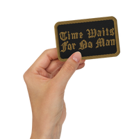 A hand holds the Mainstay Entertainment Time Waits For No Man "Logo" Embroidered Patch—rectangular, black, with gold trim and ornate text reading “Time Waits For No Man.”.