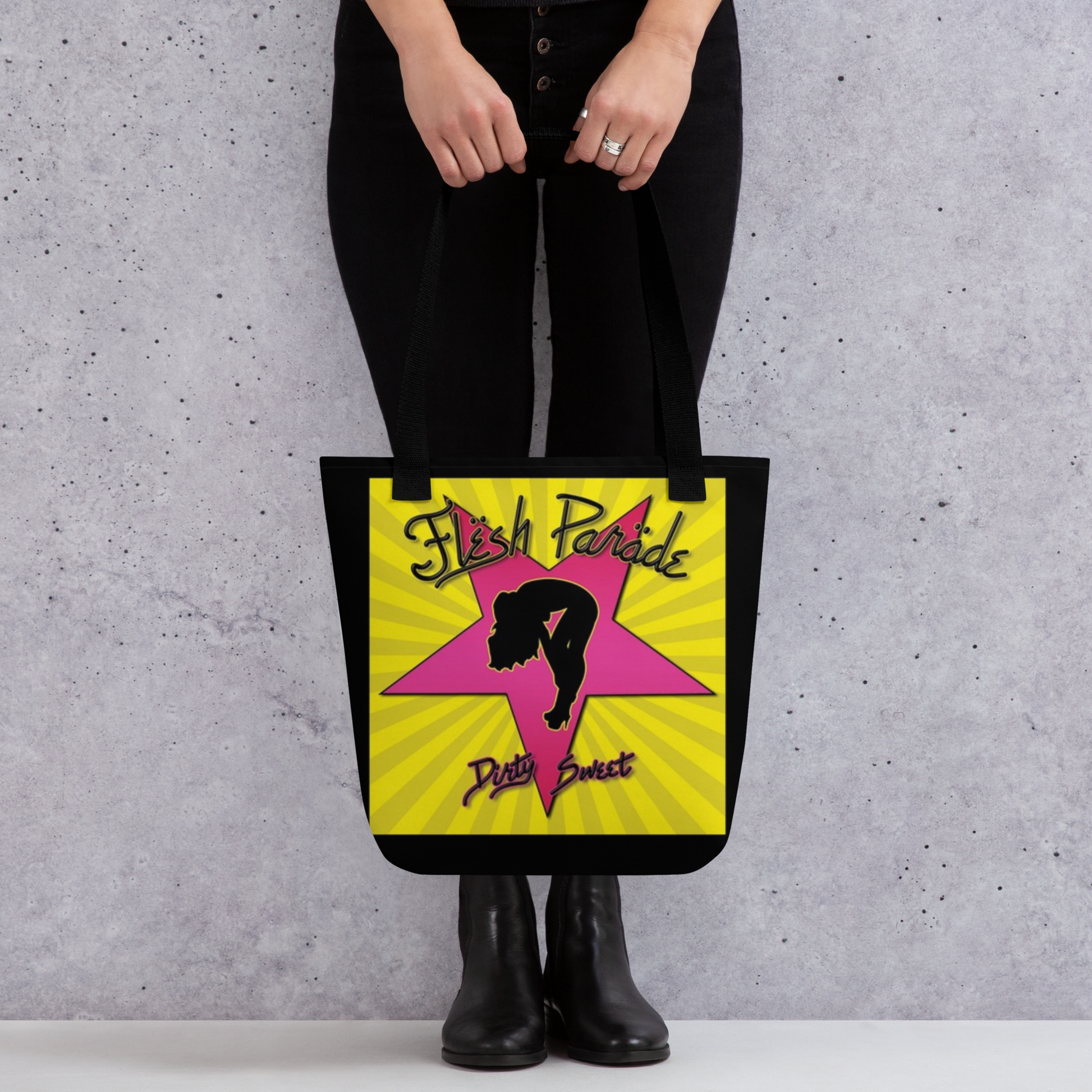A person in black pants and boots holds the Flesh Parade "Dirty Sweet" Tote Bag by Mainstay Entertainment, featuring a yellow star, pink dancer silhouette, stylized Flesh Parade and Dirty Sweet text, and bold yellow rays in the background.