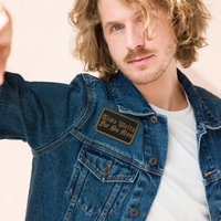 A man with wavy blond hair and a beard wears a denim jacket featuring Mainstay Entertainment's Time Waits For No Man 