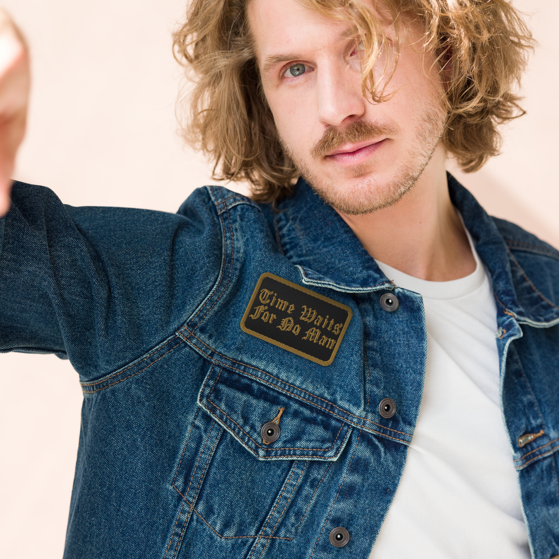 A man with wavy blond hair and a beard wears a denim jacket featuring Mainstay Entertainment's Time Waits For No Man "Logo" Embroidered Patch over a white t-shirt, looking into the camera with a neutral expression.