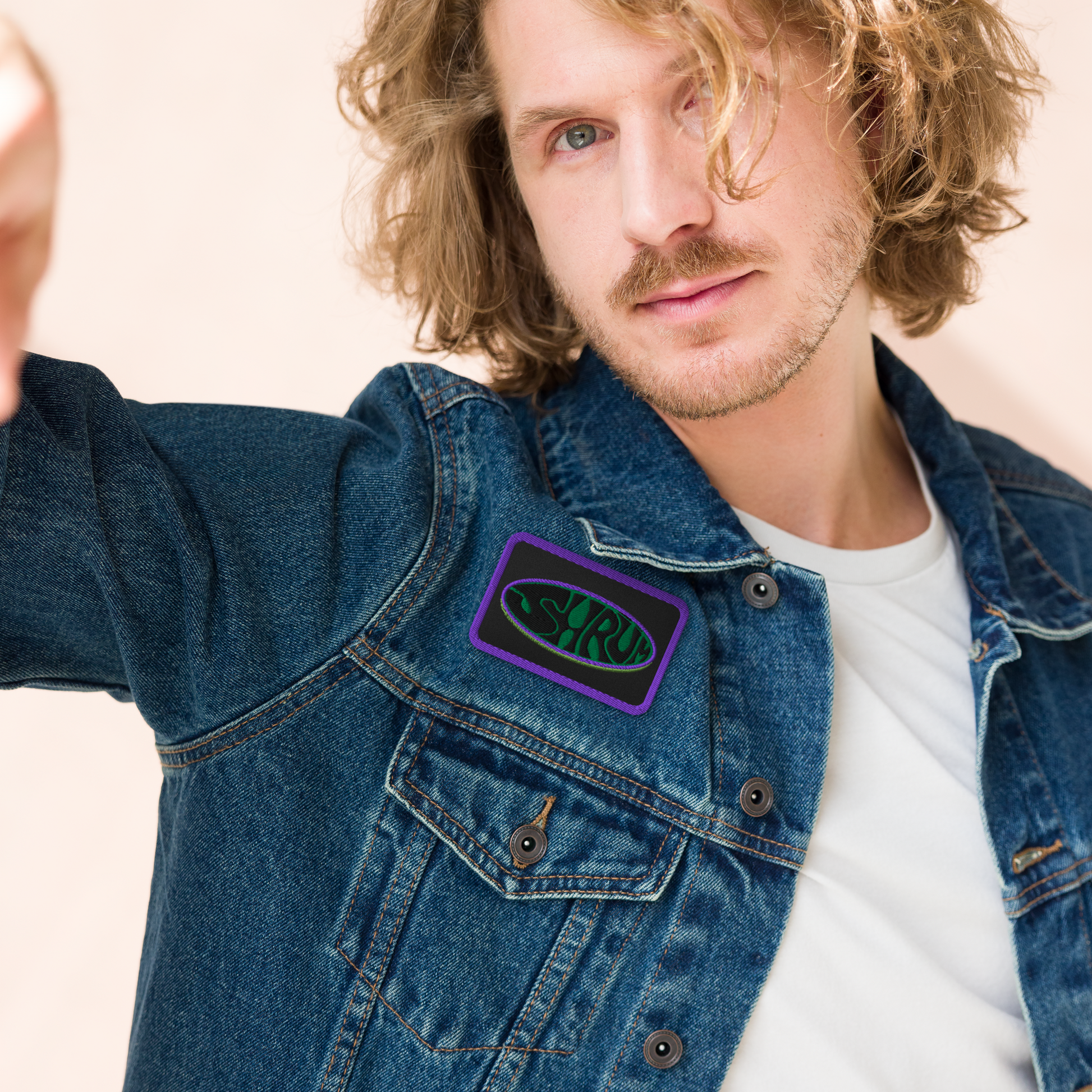 A person with wavy blonde hair in a denim jacket and white shirt displays the Mainstay Entertainment SHRÜM "Logo" Embroidered Patch, featuring "SYRUP" in green lettering, on the jacket’s chest pocket.
