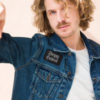 A man with wavy blonde hair and light facial hair wears a denim jacket featuring the Mainstay Entertainment Decaying Eucharist 