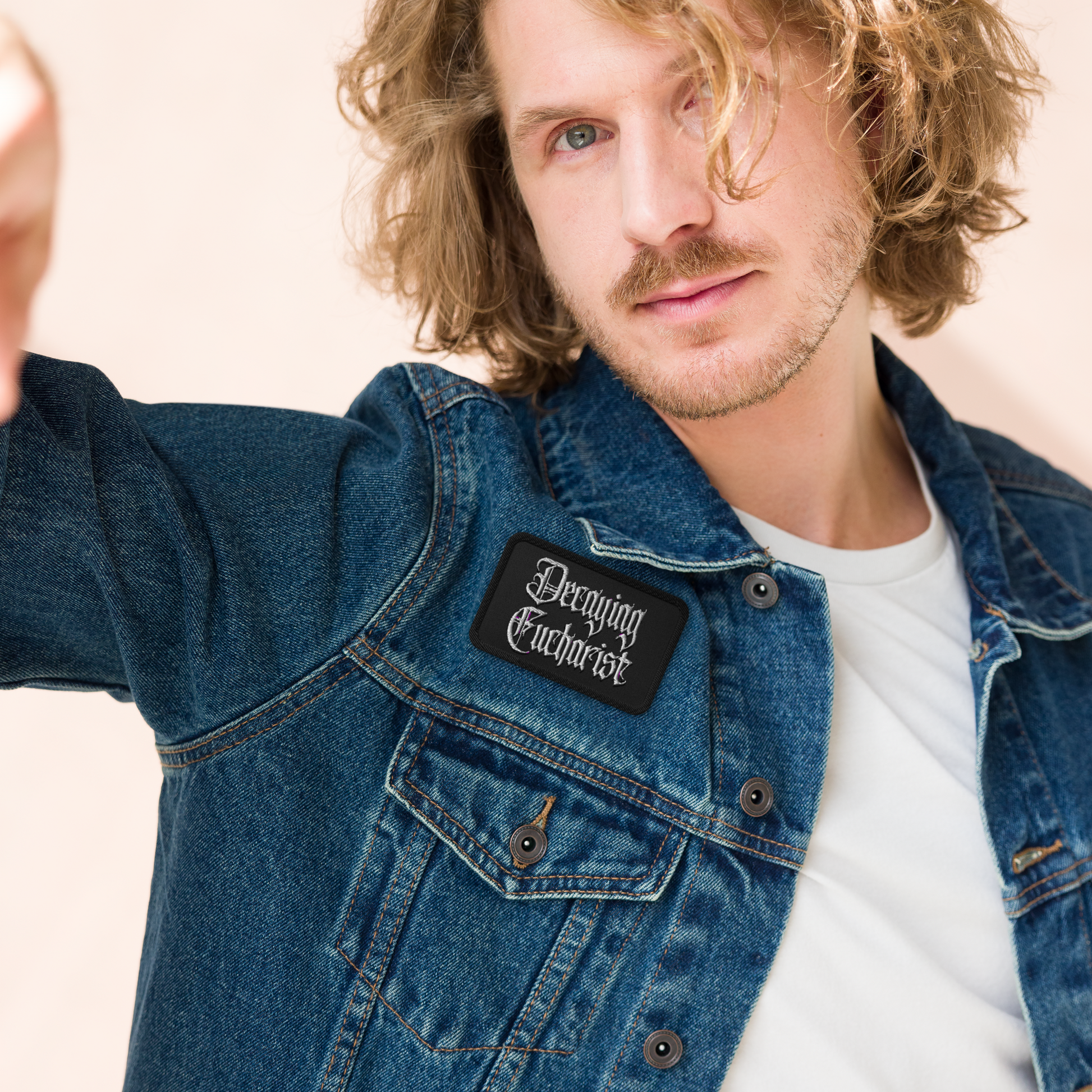 A man with wavy blonde hair and light facial hair wears a denim jacket featuring the Mainstay Entertainment Decaying Eucharist "Logo" embroidered patch in gothic script over a white T-shirt, looking at the camera.