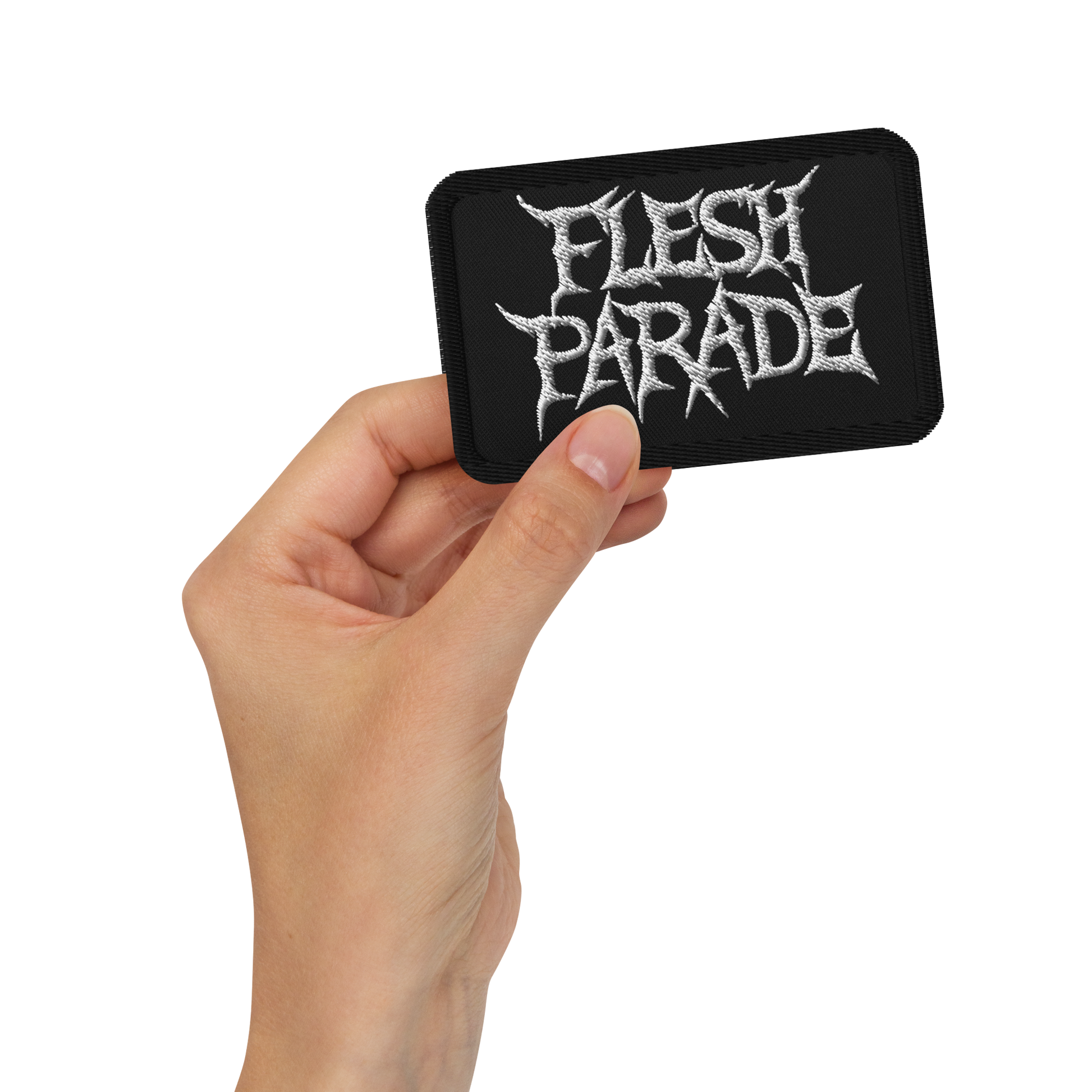 A hand holds the Mainstay Entertainment Flesh Parade "Classic Logo" embroidered patch—black, rectangular, with jagged white text—against a plain white background.
