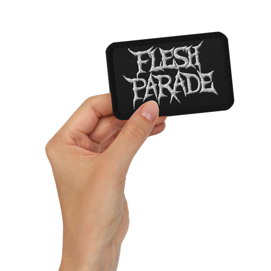 A hand holds the Mainstay Entertainment Flesh Parade "Classic Logo" embroidered patch—black, rectangular, with jagged white text—against a plain white background.