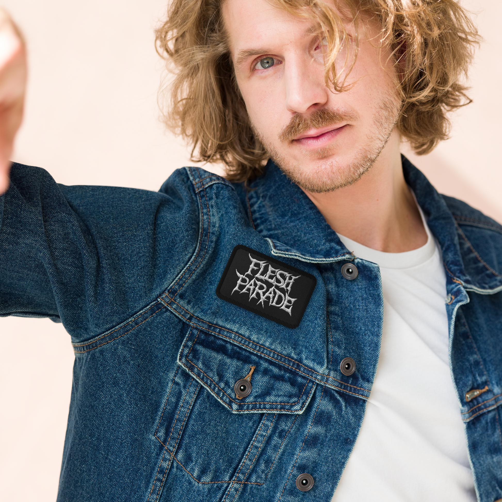 A man with wavy, shoulder-length blond hair wears a denim jacket featuring the Mainstay Entertainment Flesh Parade "Classic Logo" Embroidered Patch over a white t-shirt, looking at the camera with a neutral expression.