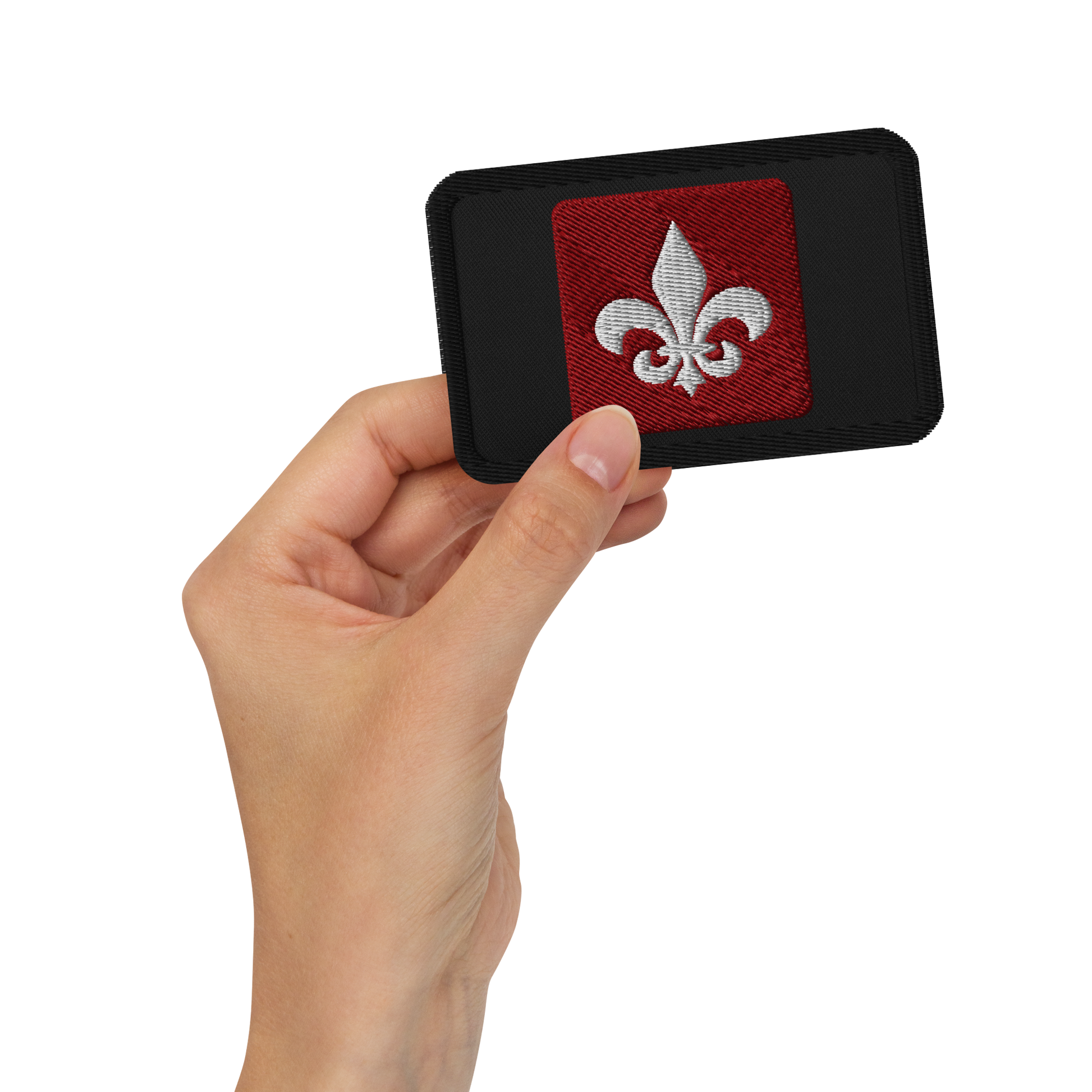 A hand holds the Mainstay Entertainment "Fleur de Lis" Embroidered Patch—a rectangular black patch with a red square center and a white fleur-de-lis symbol.