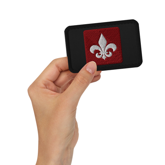 A hand holds the Mainstay Entertainment "Fleur de Lis" Embroidered Patch—a rectangular black patch with a red square center and a white fleur-de-lis symbol.