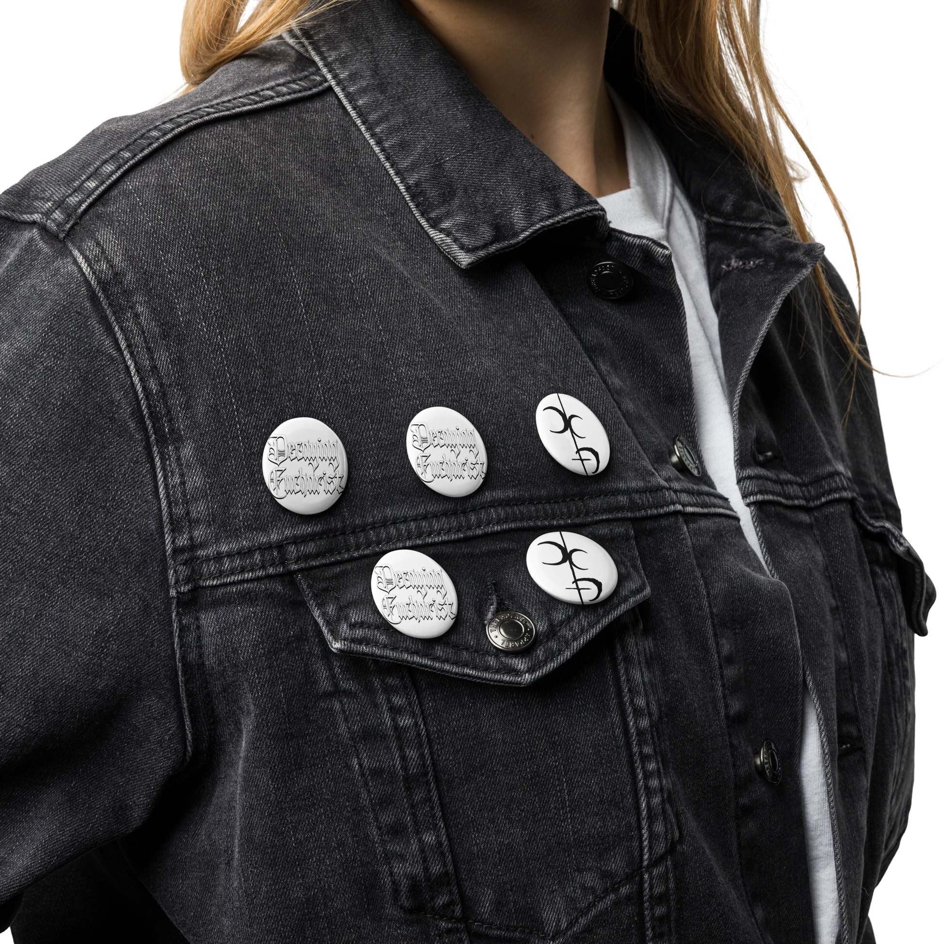 A person wears a black denim jacket and white shirt, displaying Mainstay Entertainment’s Decaying Eucharist "Logo" Set of (5) scratch and UV-resistant pin buttons with black designs and text on the jacket’s upper left side and pocket flap.