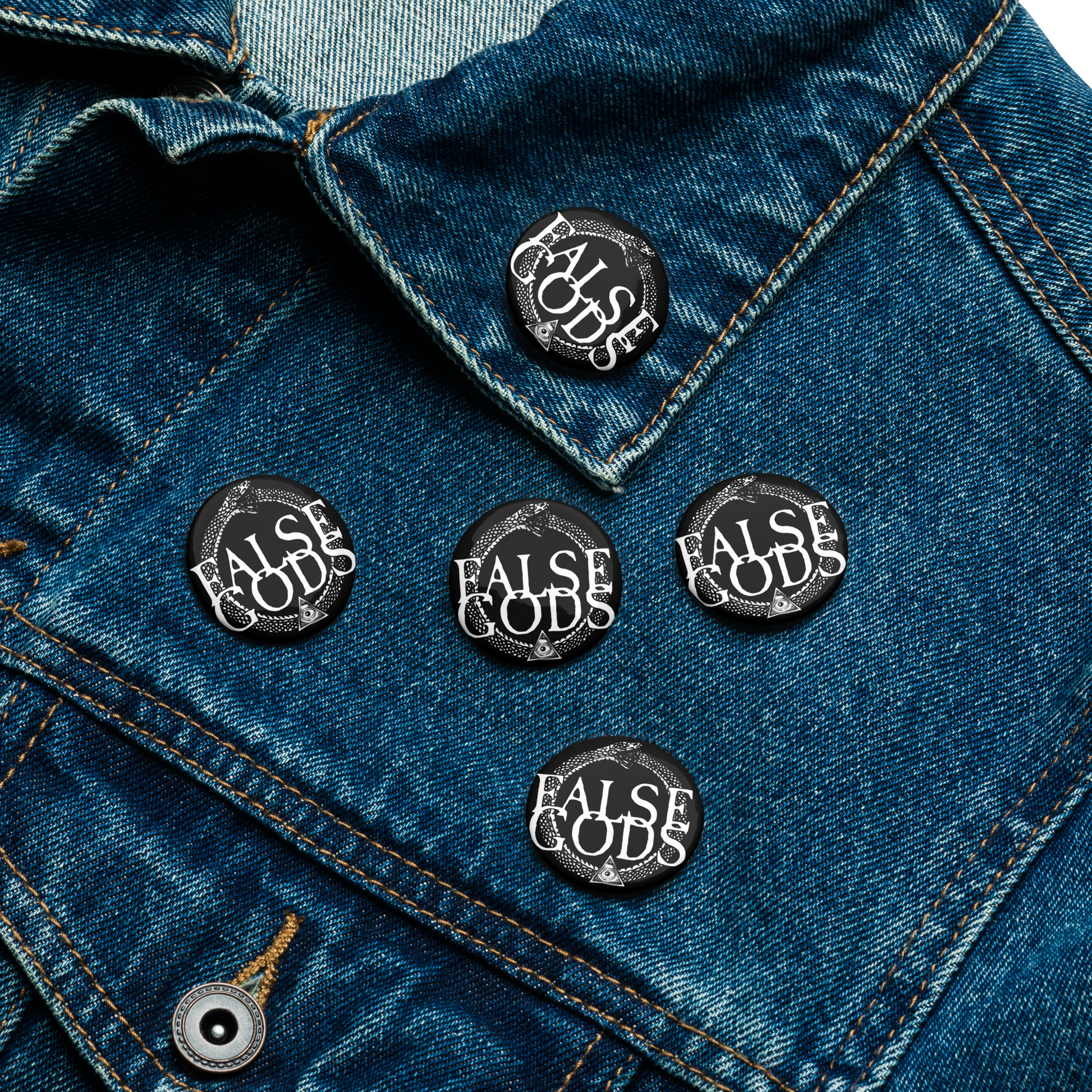 A close-up of five black False Gods "Snake Logo" Buttons by Mainstay Entertainment on a blue denim jacket lapel. Their glossy, UV-resistant finish keeps them bold and eye-catching. Set includes 5 buttons.