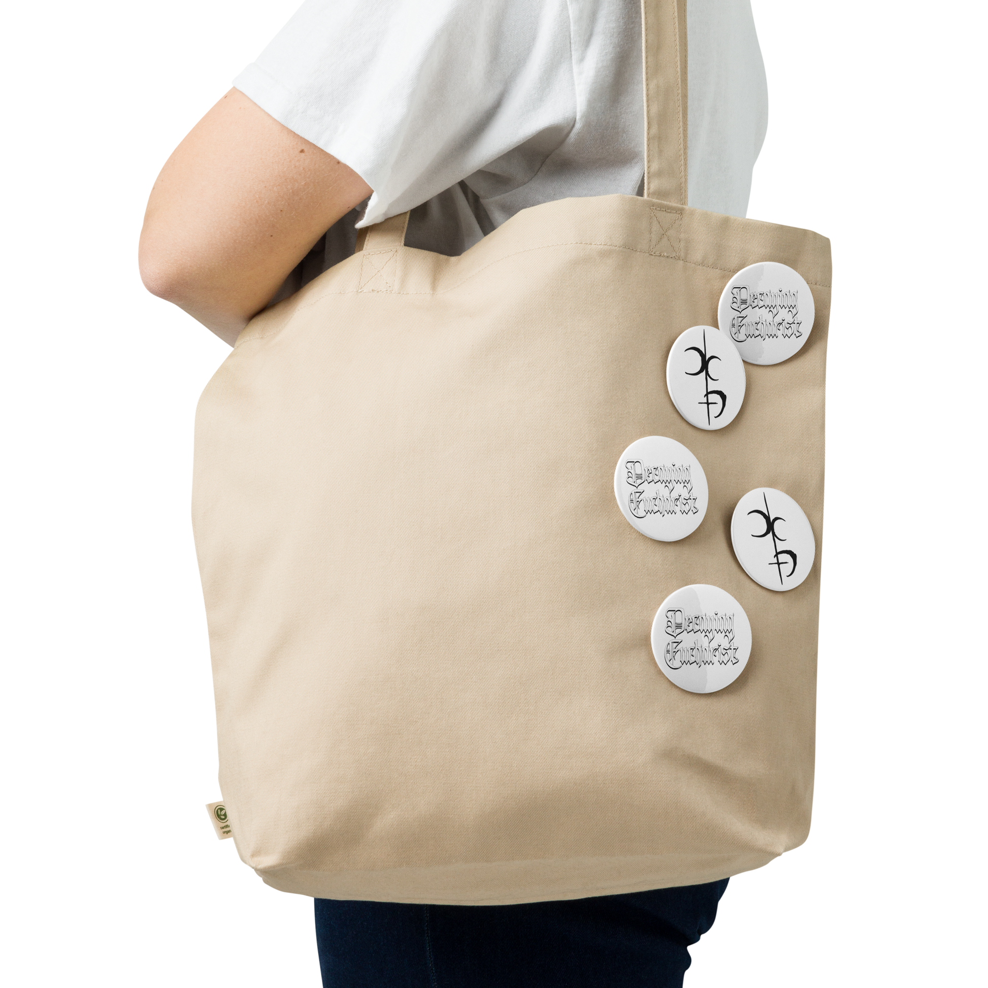 A person carries a beige tote bag decorated with Mainstay Entertainment’s Decaying Eucharist "Logo" Set of 5 buttons, each with black handwritten-style designs, attached to the side, while wearing a white shirt.