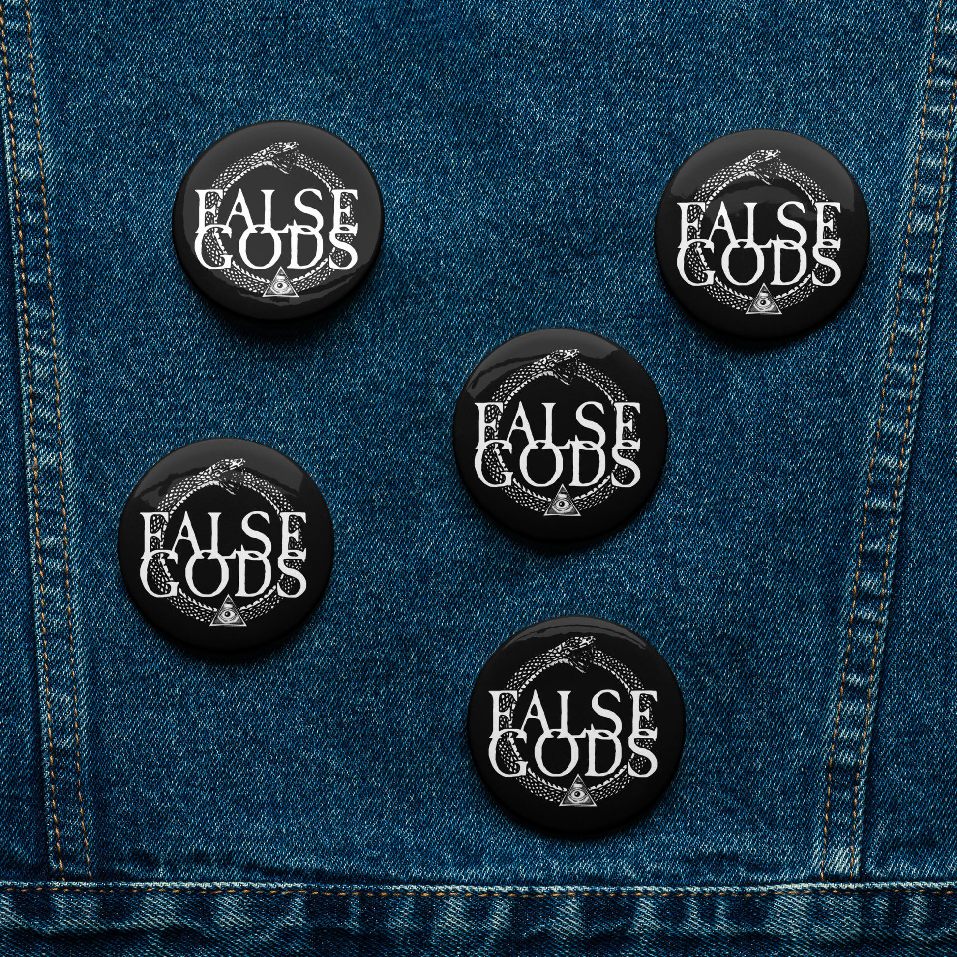 The Mainstay Entertainment False Gods "Snake Logo" Set of 5 glossy buttons featuring a snake design and the words FALSE GODS is displayed on blue denim fabric.