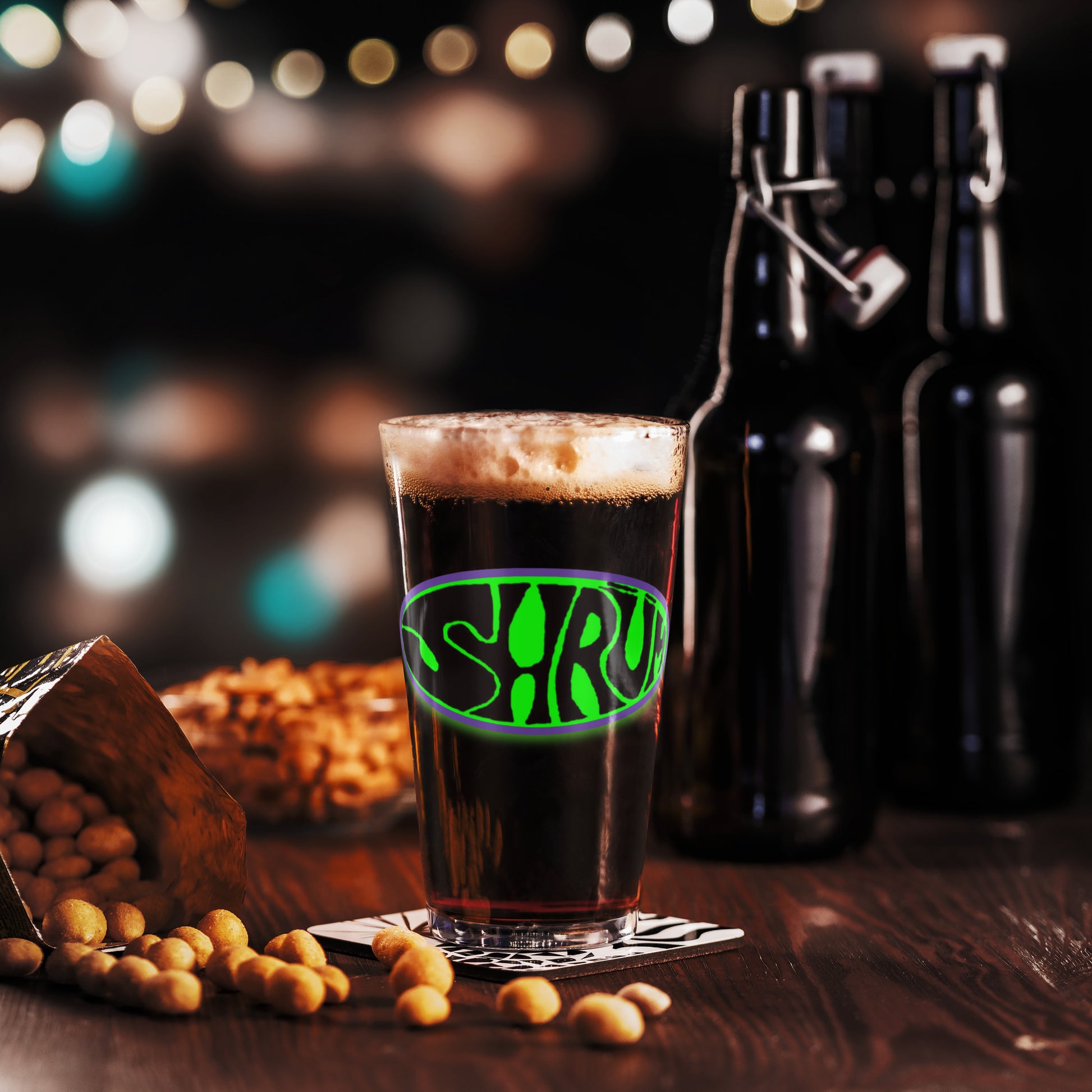 A SHRÜM "Logo" Pint Glass by Mainstay Entertainment sits on a table with peanuts, flanked by two dark bottles and festive blurred lights in the background, creating a warm, inviting scene.