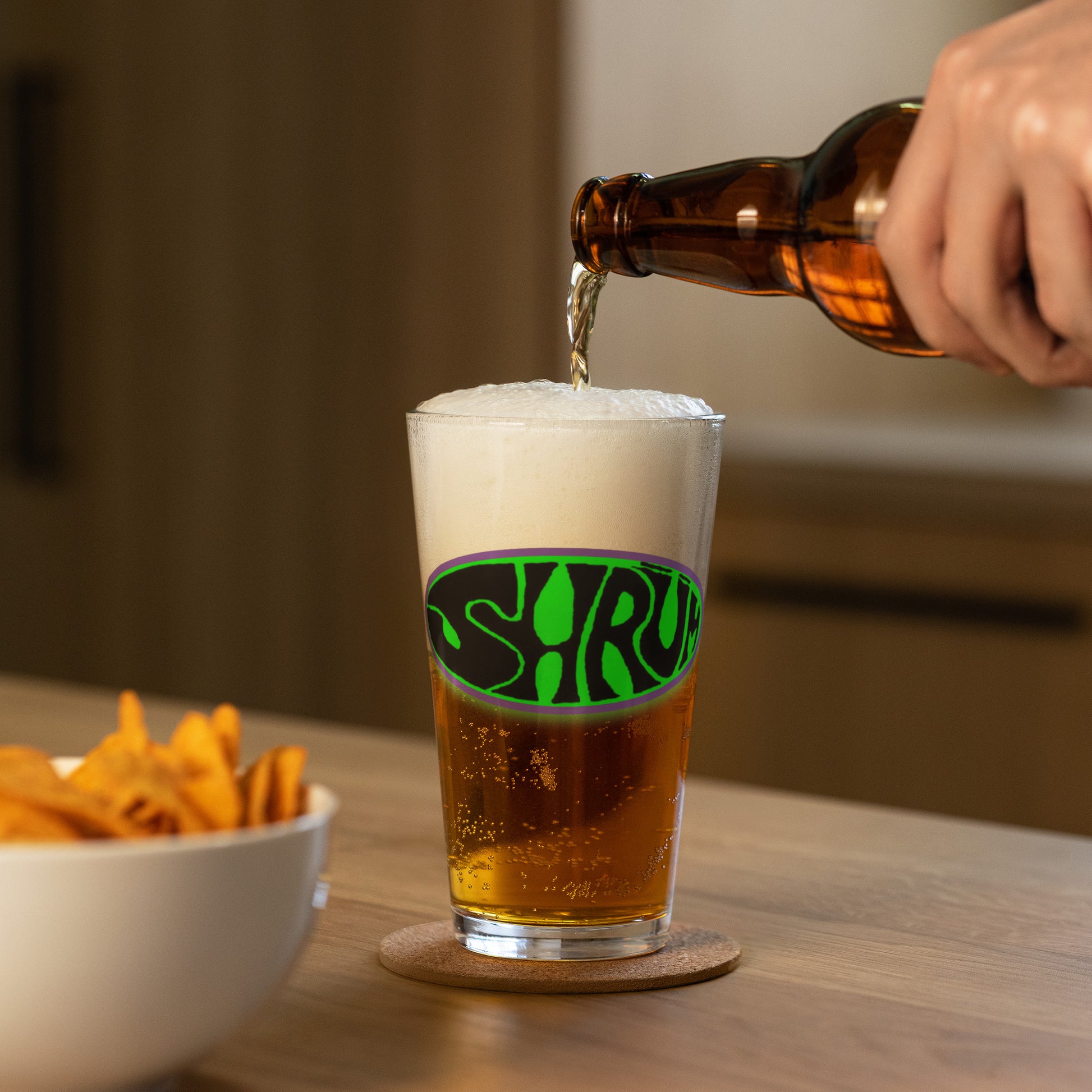 A hand pours beer from a brown bottle into a foamy SHRÜM "Logo" Pint Glass by Mainstay Entertainment, among assorted glassware and a snack bowl on a wooden table.