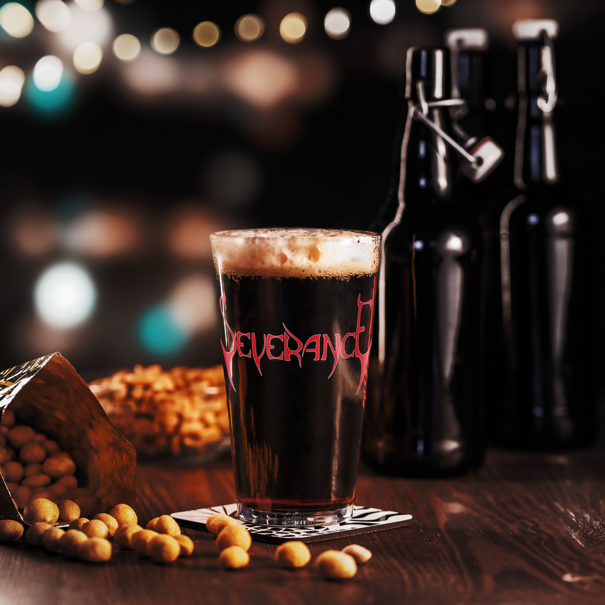 A Severance "Logo" Pint Glass by Mainstay Entertainment sits on a wooden table with peanuts and two dark bottles nearby, blurred lights glowing in the background.
