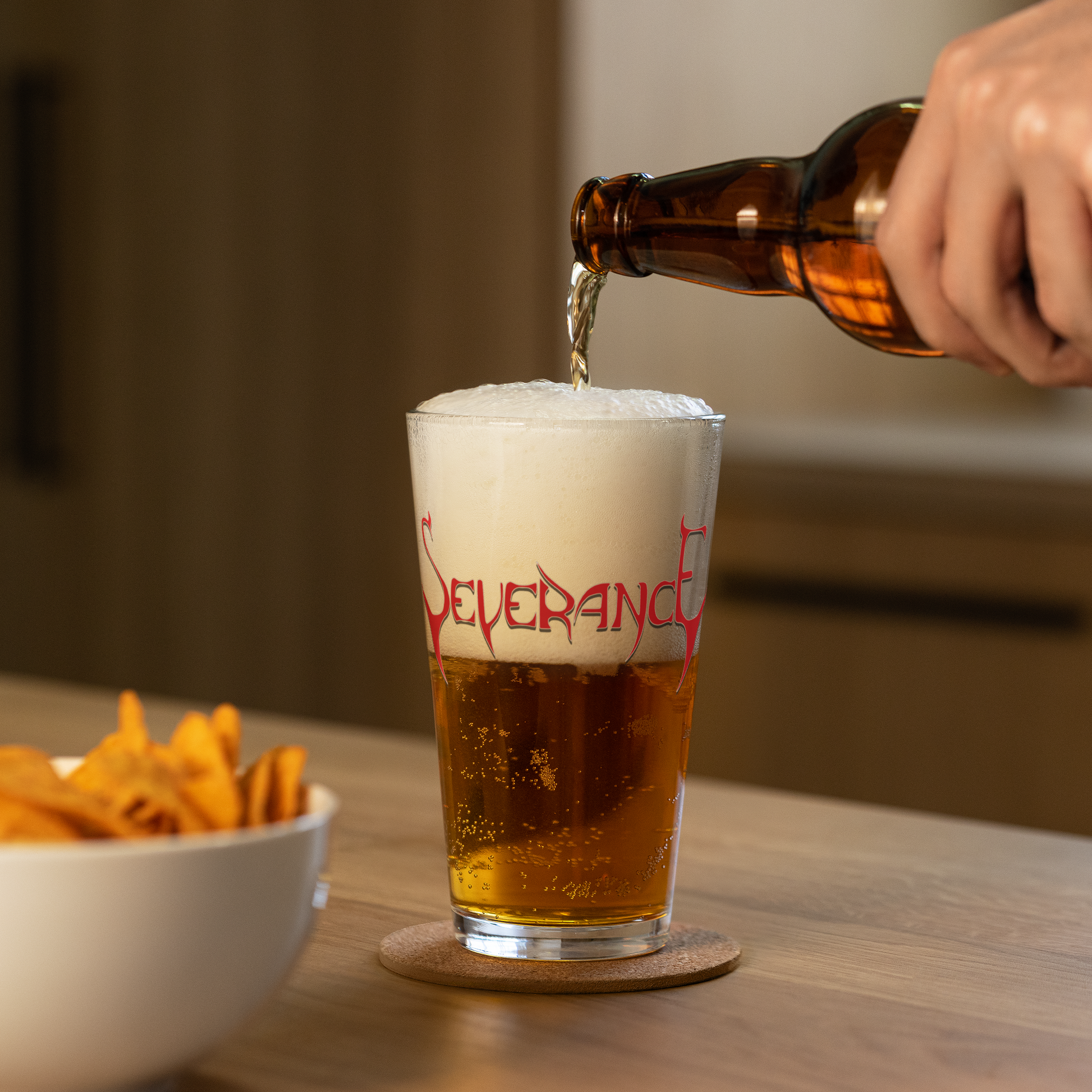 A hand pours beer from a brown bottle into a 16 oz Severance "Logo" Pint Glass by Mainstay Entertainment, creating a large foamy head, with a bowl of chips nearby on a wooden table.