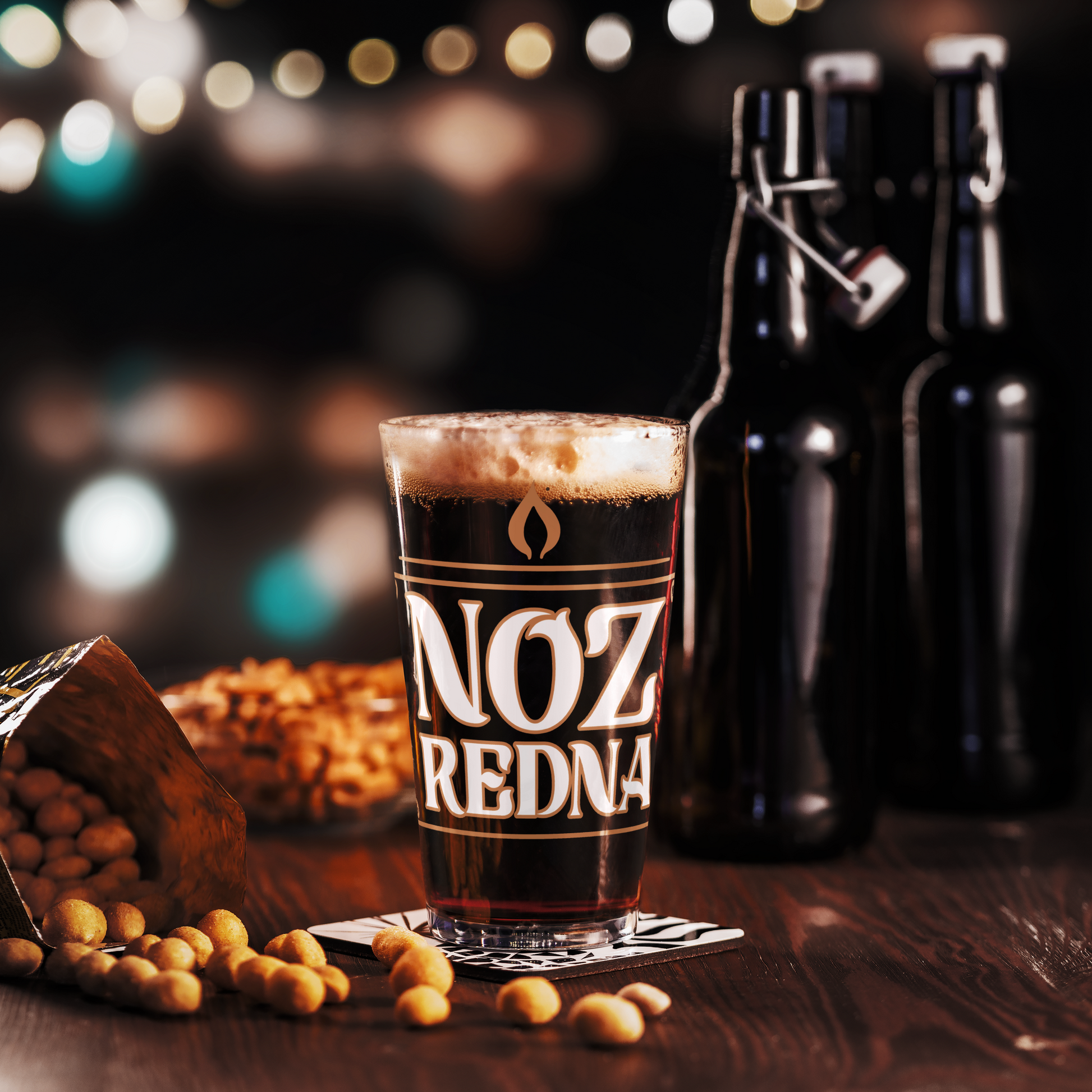 A 16 oz Noz Redna "Candle Logo" Pint Glass by Mainstay Entertainment sits on a coaster next to peanuts and two swing-top bottles on a wooden table, with blurred lights in the background.