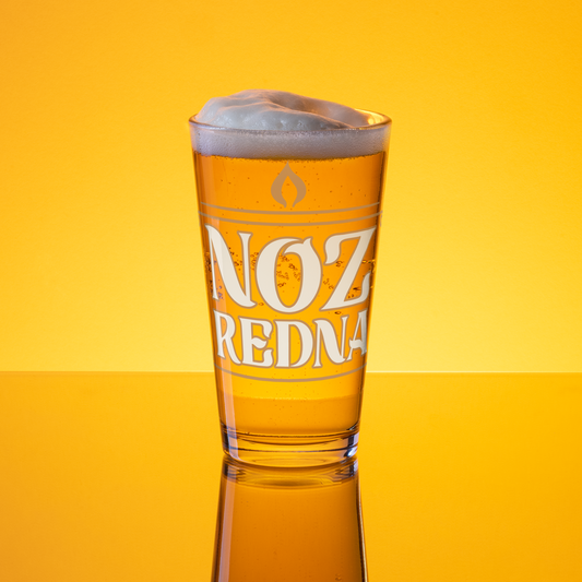 A 16 oz Noz Redna "Candle Logo" Pint Glass from Mainstay Entertainment, filled with amber beer and a foamy head, rests on a reflective surface against a bright yellow background.