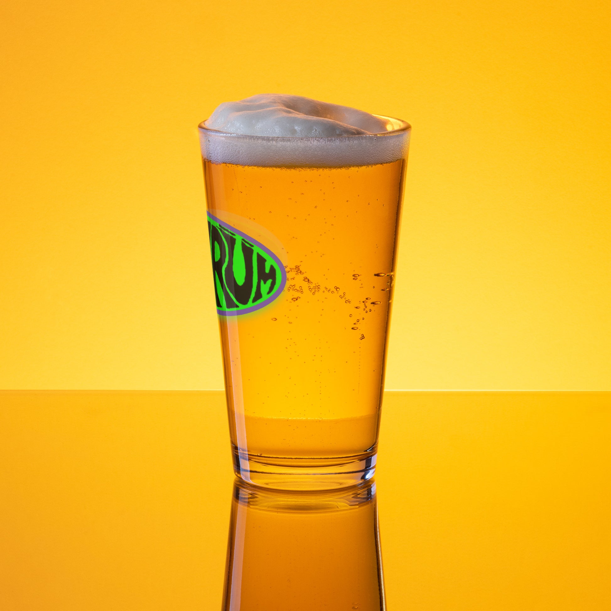 A 16 oz SHRÜM "Logo" Pint Glass by Mainstay Entertainment, filled with golden beer and a foamy head, sits on a reflective surface against a yellow-orange background. The glass displays a green and yellow logo.