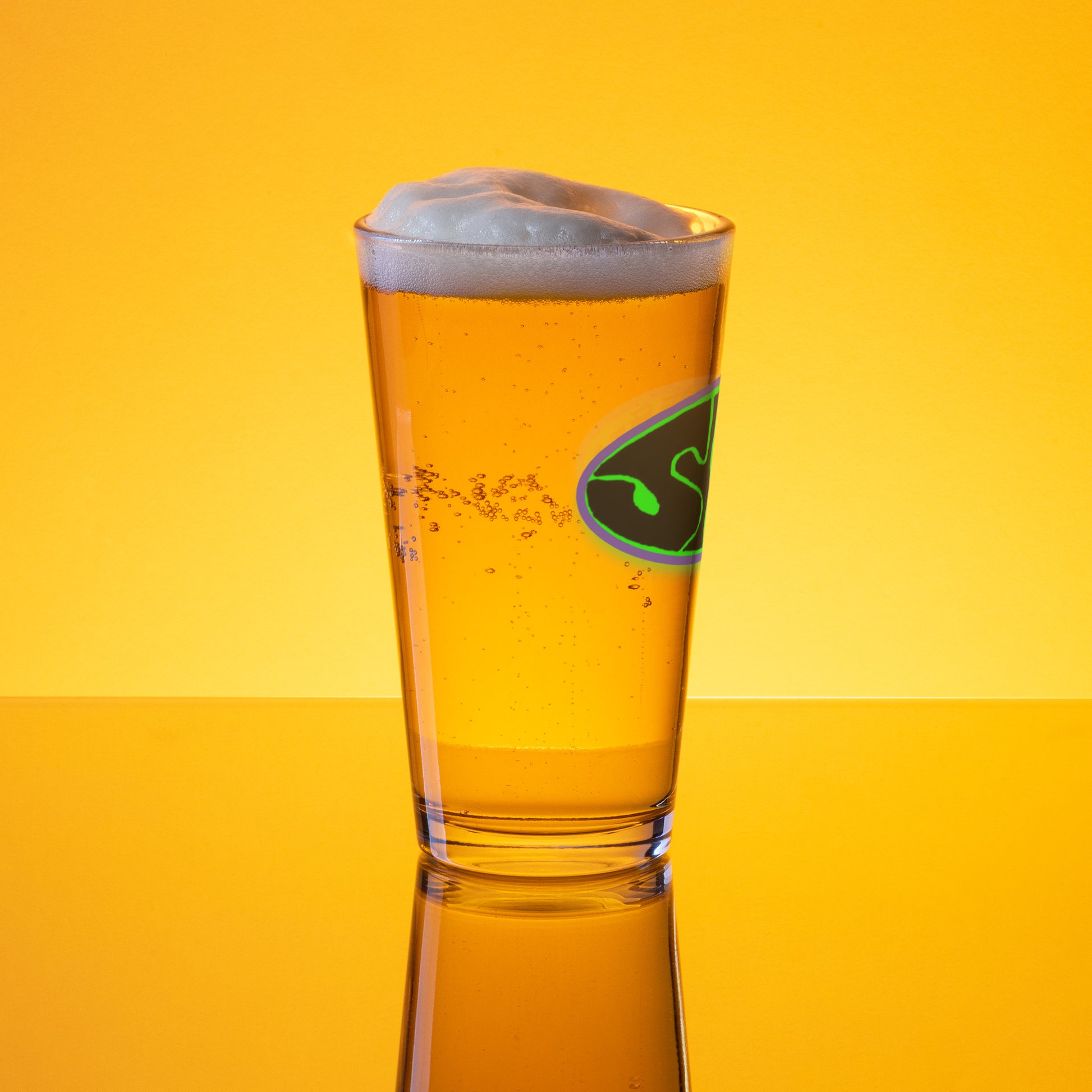 A SHRÜM "Logo" Pint Glass by Mainstay Entertainment, filled with golden beer and topped with a frothy head, rests on a reflective surface against a vibrant yellow-orange background.