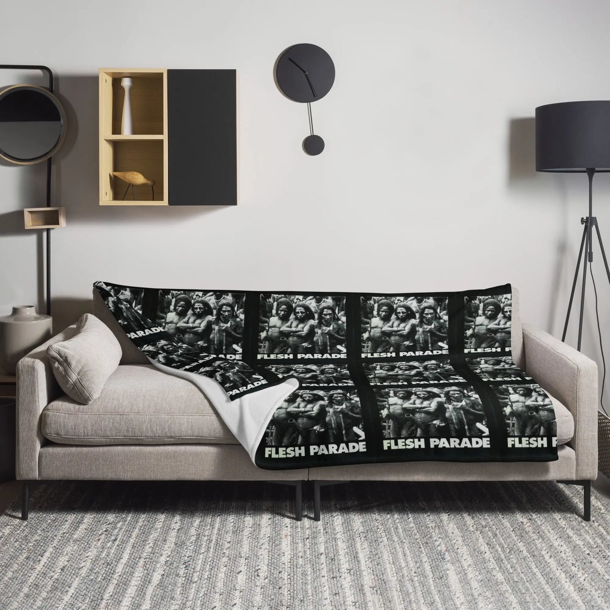 A modern living room features a beige sofa adorned with the Flesh Parade "Kill Whitey" Throw Blanket, displaying repeated black-and-white images and text. A gray rug, black floor lamp, and wall shelves complete the space.