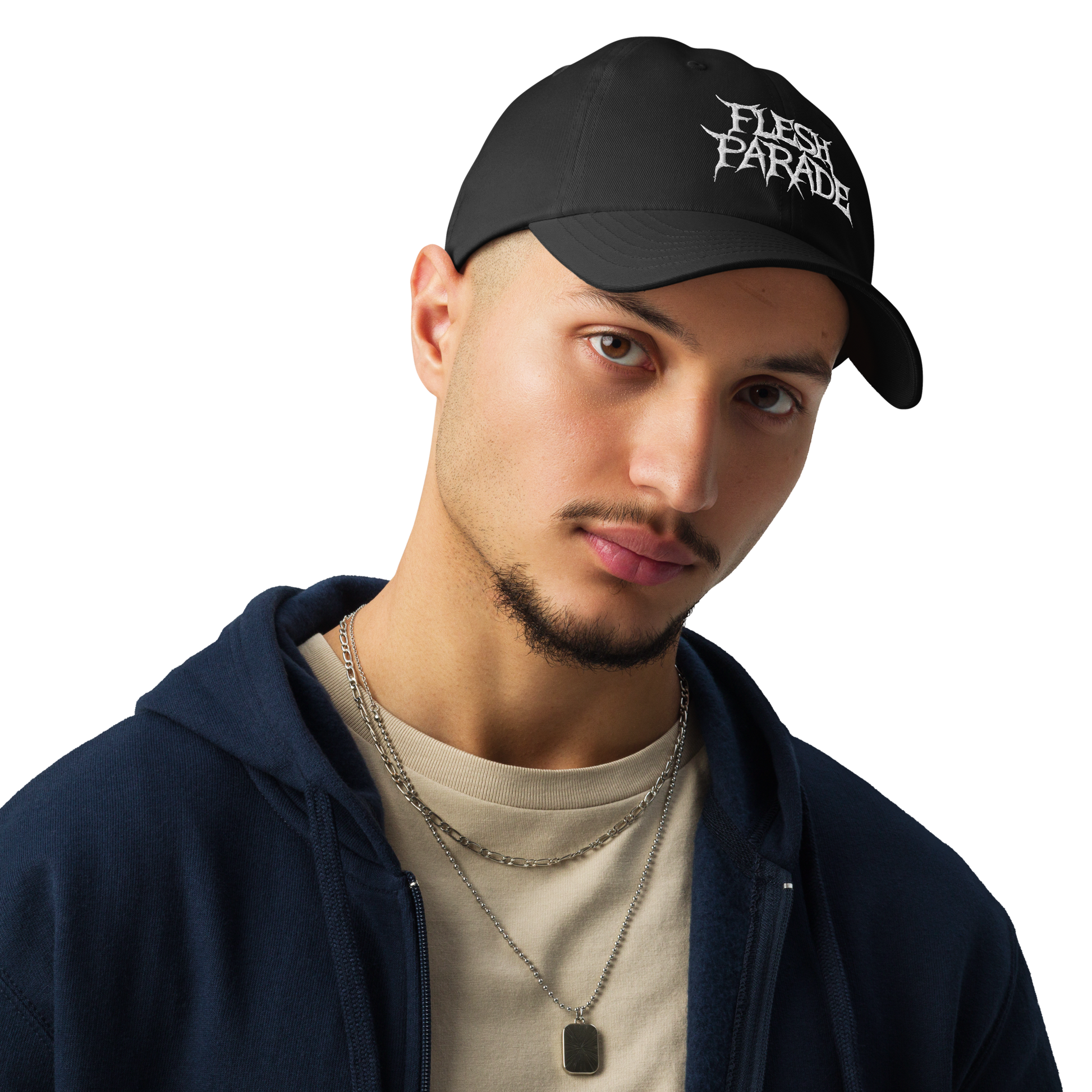 A young man wears the Flesh Parade "Embroidered Logo" Under Armour® Hat in black, paired with a beige t-shirt, blue zip-up hoodie, and layered silver necklaces as he looks at the camera against a plain backdrop.